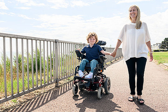 Ein Kind fährt auf einem Parkweg in einem Rollstuhl. Es hält die Hand einer Frau neben ihm.