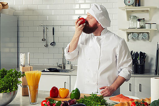 Ein Koch steht vor einer Küchenarbeitsfläche voller Gemüse und riecht an einer Tomate.