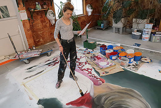 Ein riesiges, halbfertiges Bild liegt auf dem Boden. Eine junge Frau steht darauf und malt mit einem Pinsel, dessen Stiel ihr bis zur Hüfte reicht.