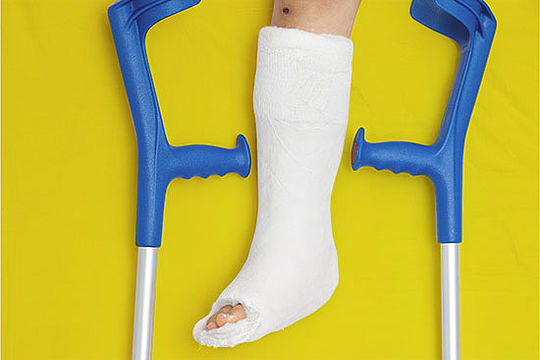 Ein Bein in Gips vor einfarbigem Hintergrund, links und rechts davon je eine Krücke.