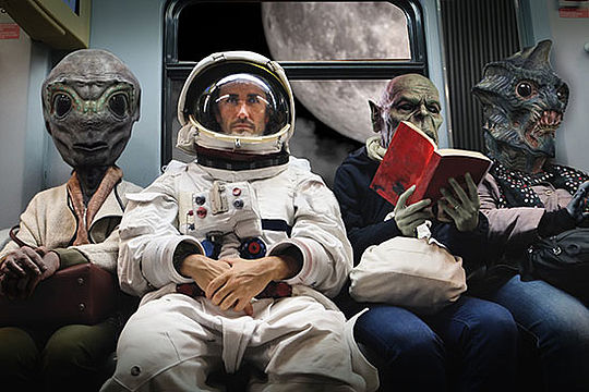 Szene aus der Zukunft: Ein Astronaut sitzt zwischen Außerirdischen auf einer Bank.