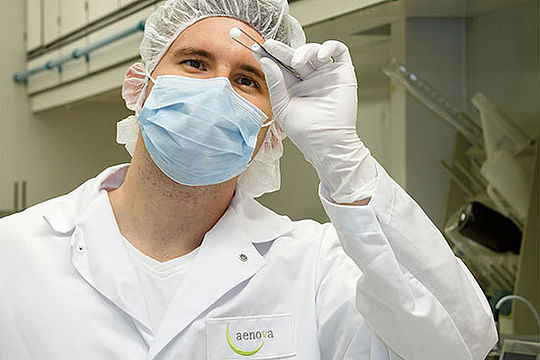 Junger Mann in Laborkleidung und mit Mundschutz hält mit einer Pinzette eine Tablette.