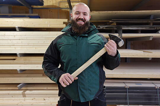 Jonas hält einen Hammer in der Hand und steht in einem Holzlager.