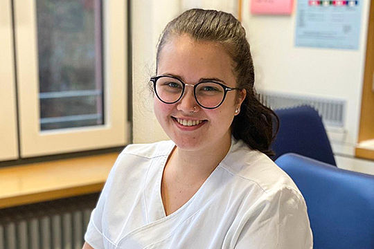 Eine junge Frau in weißer Krankenhaus-Arbeitskleidung sitzt an einem Schreibtisch und lacht in die Kamera.